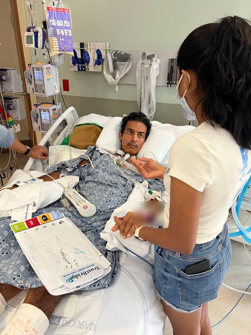 Karamat awake with a tracheostomy, his sister at the bedside, and the clipboard he used to communicate resting on the bed